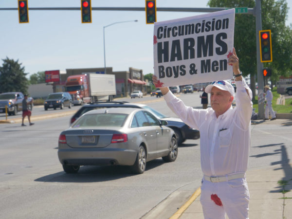 A bloodstained man holds a sign above his head – "Circumcision Harms Boys and Men"