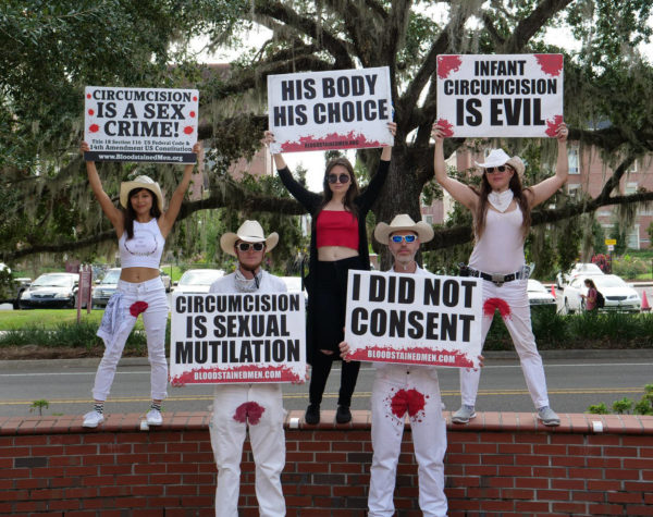 Five protesters hold signs reading "Circumcision is Sexual Mutilation", "I Did Not Consent", "Circumcision is a Sex Crime", "His Body His Choice", and "Infant Circumcision is Evil".