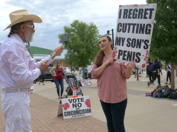 A woman holds a sign reading "I Regret Cutting My Son's Penis"