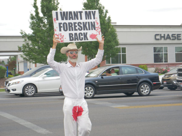 A bloodstained man holds a sign above his head – "I Want My Foreskin Back"