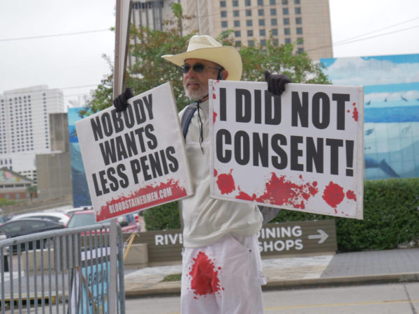 A bloodstained man holds two signs – "Nobody Wants Less Penis" and "I Did Not Consent"