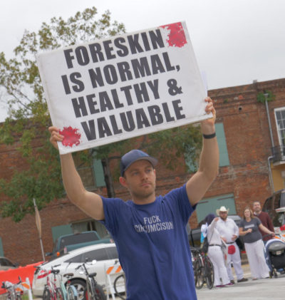 A man holds a sign reading "Foreskin is Normal, Healthy, and Valuable"