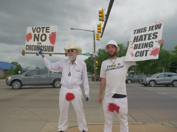 Two bloodstained men in Colorado hold protest signs – "Vote No on Circumcision" and "This Jew Hates Being Cut"