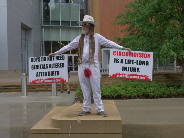 A bloodstained protester holds two signs – "Boys Do Not Need Genitals Altered After Birth" and "Circumcision is a Lifelong Injury"