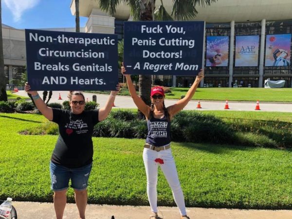 Two women hold signs reading "Fuck You, Penis Cutting Doctors! - A Regret Mom" and "Non-therapeutic Circumcision Breaks Genitals and Hearts"