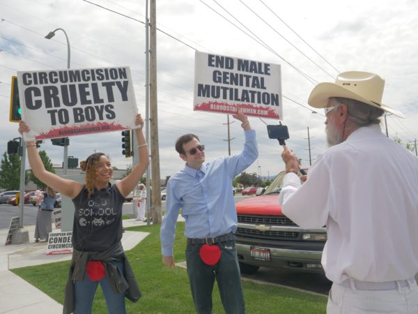 Two protesters in street clothes with felt bloodstains pinned to their crotches hold signs reading "Circumcision Cruelty to Boys" and "End Male Genital Mutilation"